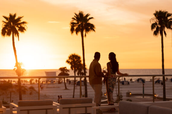 A couple stands on a terrace at sunset, holding drinks and looking out over a beach with palm trees, people, and the ocean in the background. The sky is orange and golden.