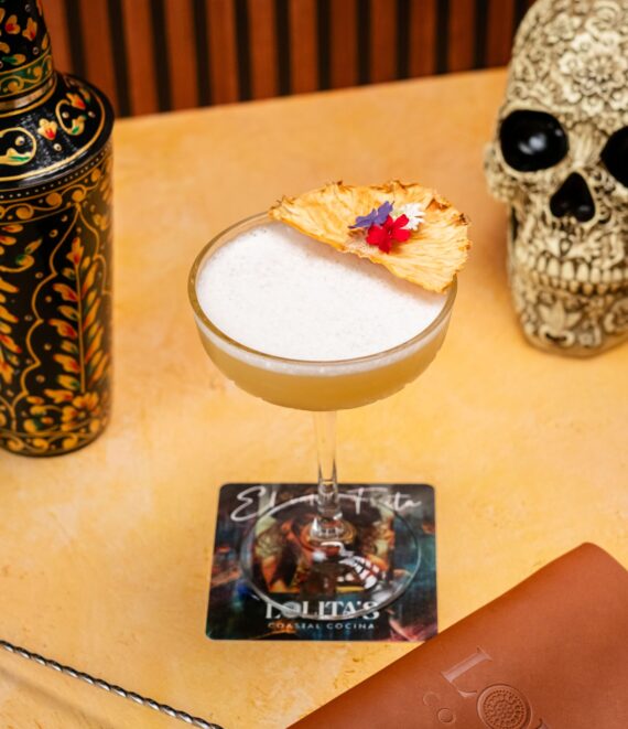 A cocktail with frothy top garnished with a dried pineapple slice and tiny flowers, sits on a Lolitas coaster. Nearby are a decorative shaker, a skull ornament, a brown menu, and a metal bar spoon.
