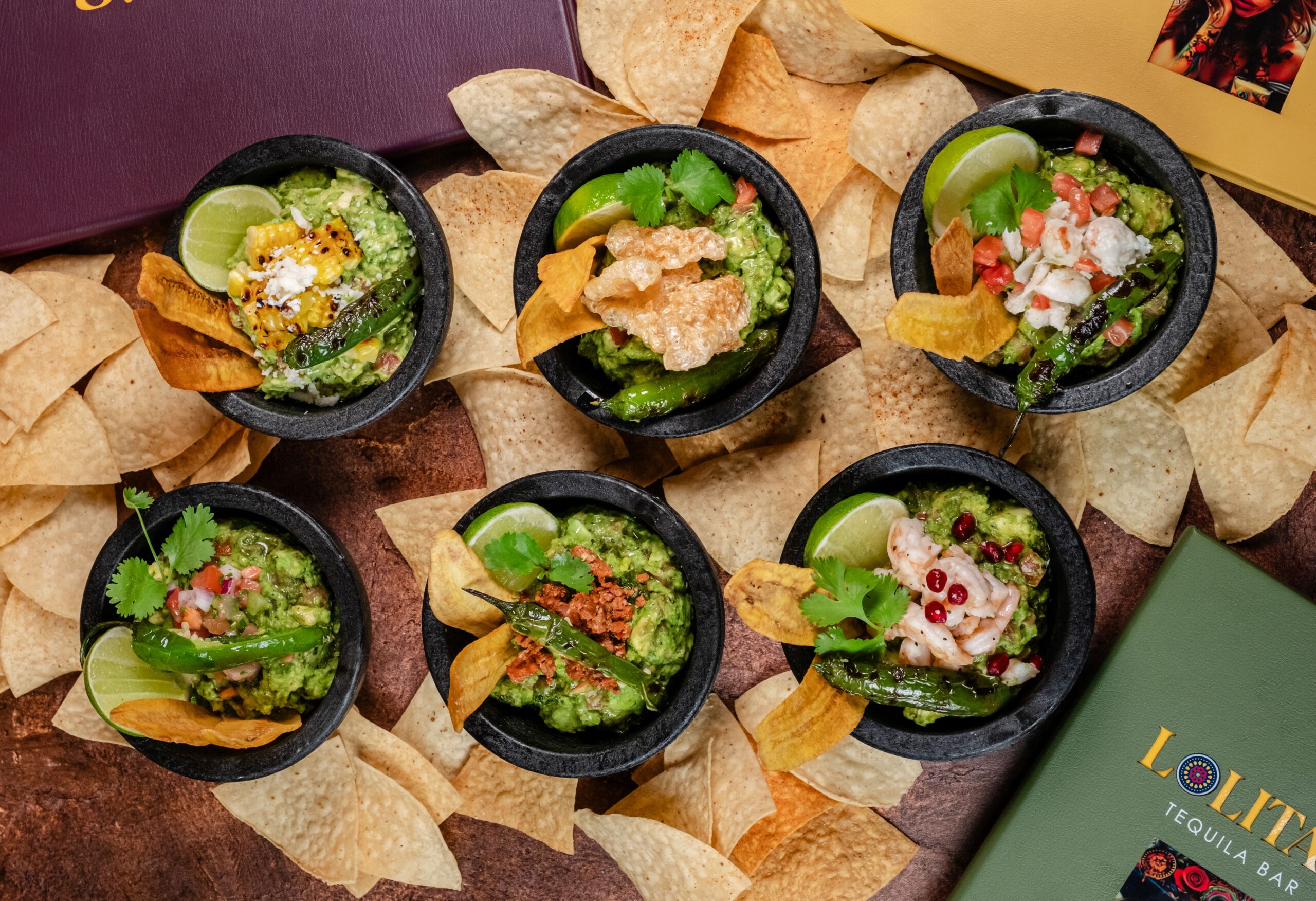 Six small bowls of guacamole with various toppings and garnishes, surrounded by tortilla chips and colorful restaurant menus on a brown surface.