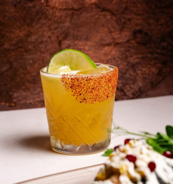 A cocktail garnished with a lime slice and chili powder on the rim sits on a table. A blurred dish with sauce and pomegranate seeds is in the foreground.
