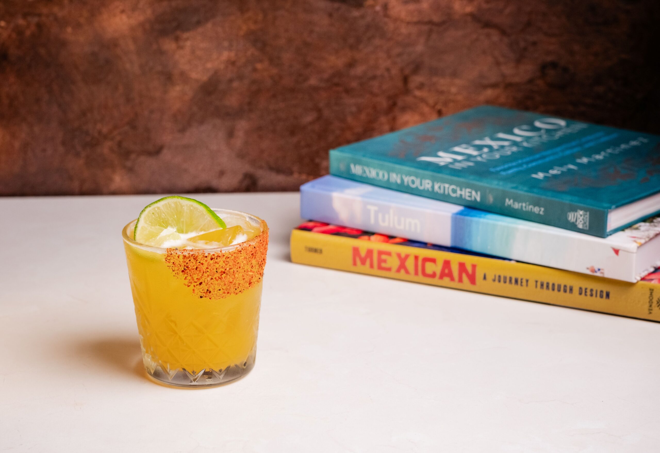 A yellow cocktail garnished with a lime wheel and chili powder sits on a white surface. Behind it are three colorful books stacked, with MEXICAN and Tulum visible on the spines. The background is brown.