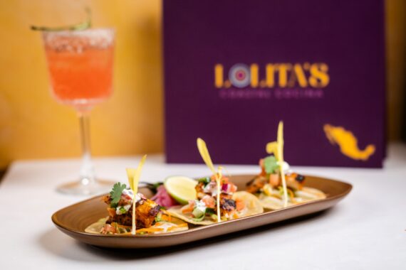 A plate of three tacos garnished with herbs and lime, next to a pink cocktail in a glass with a salted rim. A purple menu reading LOLITAS is in the background.