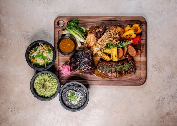 A wooden tray with grilled meats, vegetables, and sauce, served with three bowls containing salad, green rice, and black beans on a light surface.