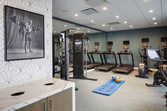 A modern gym with treadmills, stationary bikes, a weight machine, and a blue mat with kettlebells and dumbbells. A framed vintage photo of weightlifters hangs on a patterned wall. The space has bright lighting and mirrors.