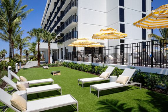 Chairs with cushions and yellow-striped umbrellas on a grassy outdoor area beside a modern white hotel, with palm trees and a cornhole game set up, under a sunny blue sky.