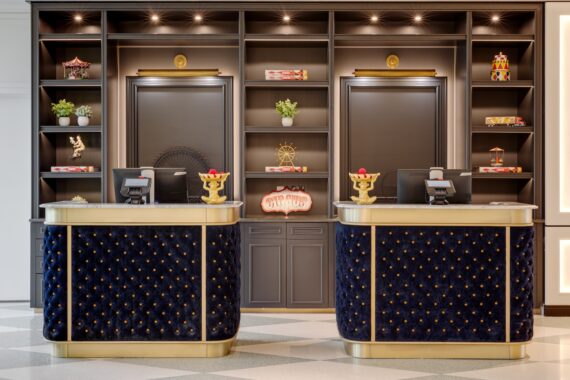 Hotel reception area with two navy and gold desks, shelving behind displaying decorative items like plants, carousel and ferris wheel models, and neon step right up sign, creating a luxurious and playful atmosphere.
