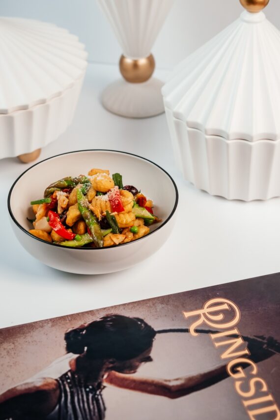 A bowl of stir-fried vegetables and chicken is placed on a white surface, surrounded by white decorative lamps. In the foreground, there is a magazine or book titled Ringside with a vintage-style cover image.