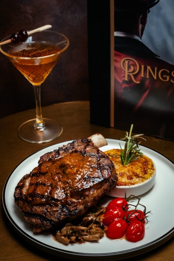 A grilled steak with sauce is served on a white plate with roasted tomatoes, mushrooms, and a baked potato side. A cocktail and a book with a partially visible cover are in the background.