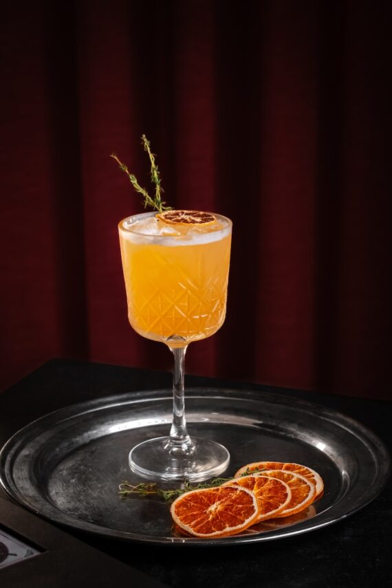 A yellow-orange cocktail in an elegant etched glass, garnished with a dried orange slice and a sprig of thyme, sits on a metal tray with additional dried orange slices against a dark red background.