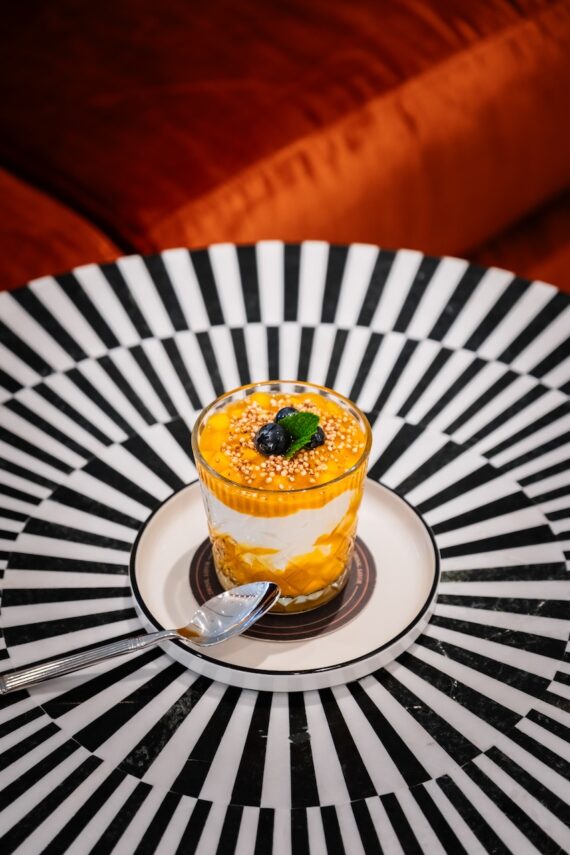 A glass dessert with layers of yellow mango and white cream, topped with blueberries, round sprinkles, and a mint leaf, sits on a striped black-and-white table with a spoon beside it.