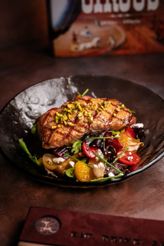 A grilled chicken breast topped with chopped pistachios sits on a bed of mixed salad greens, cherry tomatoes, and orange segments, served in a dark bowl on a brown table.