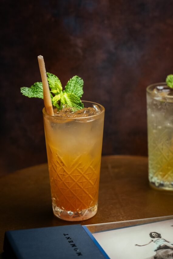 A tall glass filled with an amber-colored drink, garnished with fresh mint and a straw, sits on a brown surface next to a book and another partially visible drink.