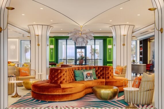 A stylish, modern hotel lobby with a curved orange velvet sofa, colorful pillows, gold coffee tables, white columns, and a large feather chandelier. Floor-to-ceiling windows reveal palm trees and ocean views in the background.