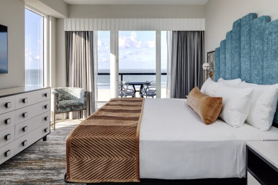 A modern hotel room with a large bed, teal upholstered headboard, tan throw blanket, and ocean view through floor-to-ceiling windows with a balcony and outdoor seating. A dresser and chair are on the left.