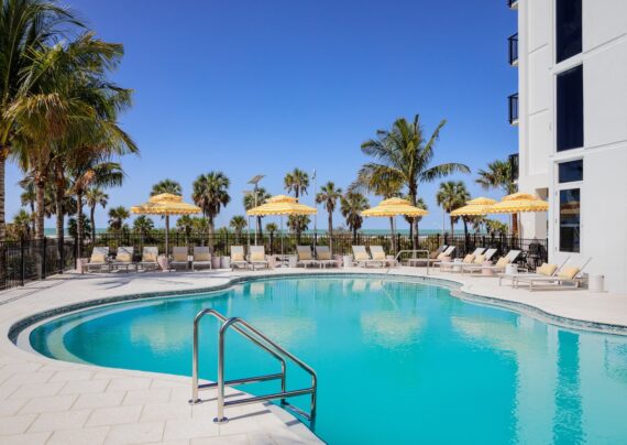 A resort-style outdoor pool with clear blue water, surrounded by lounge chairs and yellow-striped umbrellas, palm trees, and a view of the ocean under a bright blue sky.