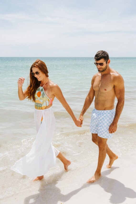 A woman and a man hold hands while walking barefoot along a sunny beach. The woman wears sunglasses, a colorful top, and a white skirt. The man wears sunglasses and blue checkered swim shorts. The sea is calm behind them.