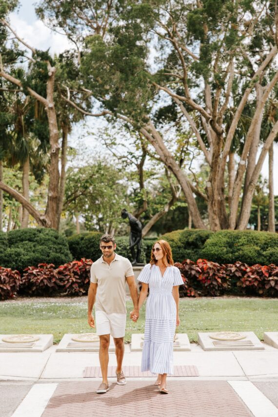 A man and woman holding hands and walking outside on a sunny day. They are both wearing sunglasses; the man is in a light polo and shorts, and the woman is in a blue-and-white striped dress. Lush trees and bushes are in the background.