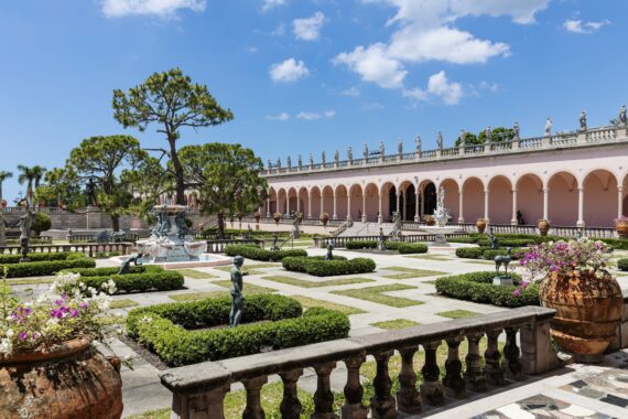 Elegant courtyard garden with manicured hedges, statues, a central fountain, and large terracotta pots under a blue sky. An arched pink building with rooftop sculptures lines the background.