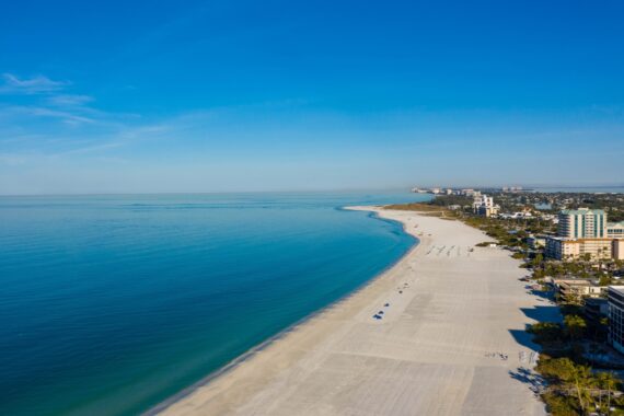 Aerial view of a wide sandy beach with calm blue water, a few umbrellas, and beachfront buildings under a clear blue sky. The coastline curves gently into the distance.
