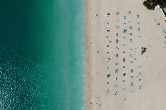 Aerial view of a sandy beach with rows of empty blue lounge chairs and umbrellas beside clear turquoise water. The shoreline curves gently, and few people are scattered across the wide, white sand.