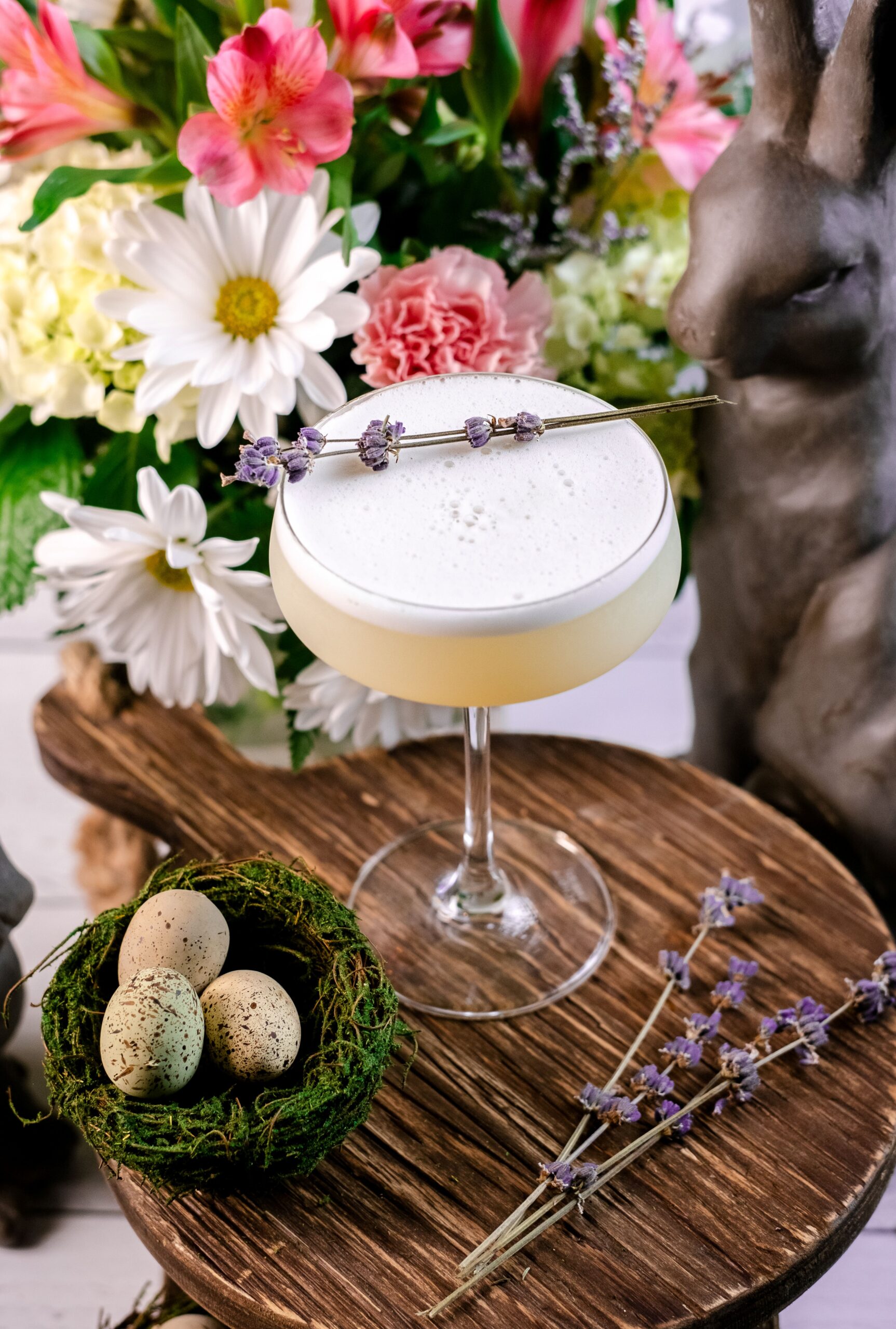 A frothy cocktail garnished with lavender rests on a wooden table beside a nest with speckled eggs and lavender sprigs, with flowers and a decorative rabbit in the background.