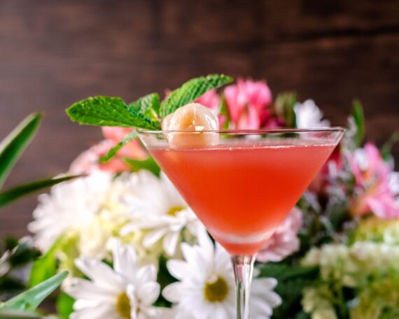 A pink cocktail in a martini glass, garnished with a lychee fruit and mint sprig, is in focus. The background is a blur of assorted colorful flowers, including white daisies and pink blooms.