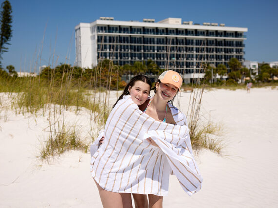 Two young people stand on a sandy beach wrapped together in a white towel with tan stripes. They smile at the camera, with a large modern hotel building and grasses in the background under a clear blue sky.