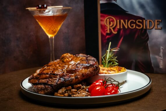 A grilled steak topped with compound butter, served with sautéed mushrooms, roasted tomatoes, and a gratin, with a cocktail and a Ringside menu in the background.