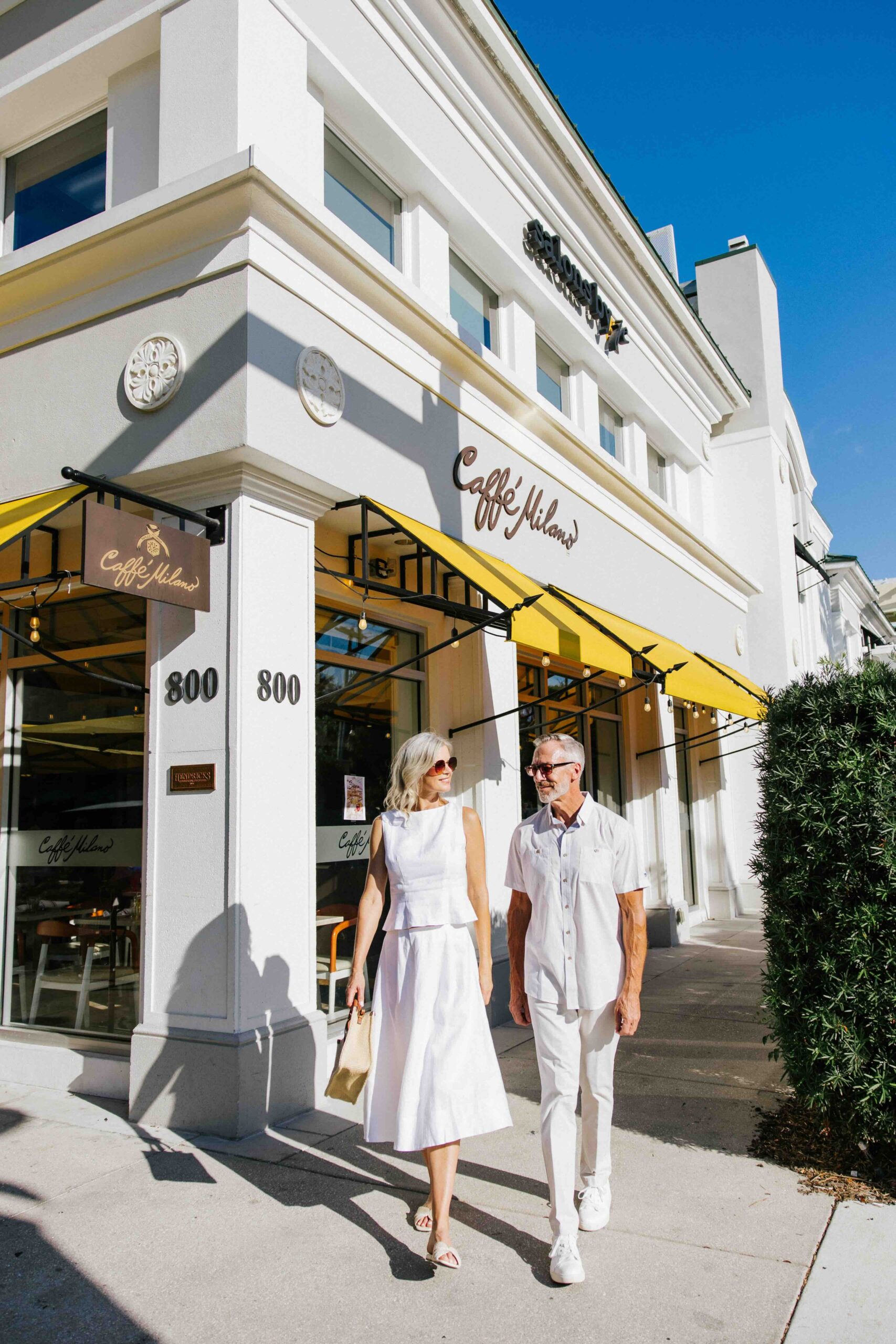 A stylish older couple wearing white summer outfits walks hand in hand on the sidewalk outside a bright café with yellow awnings on a sunny day.