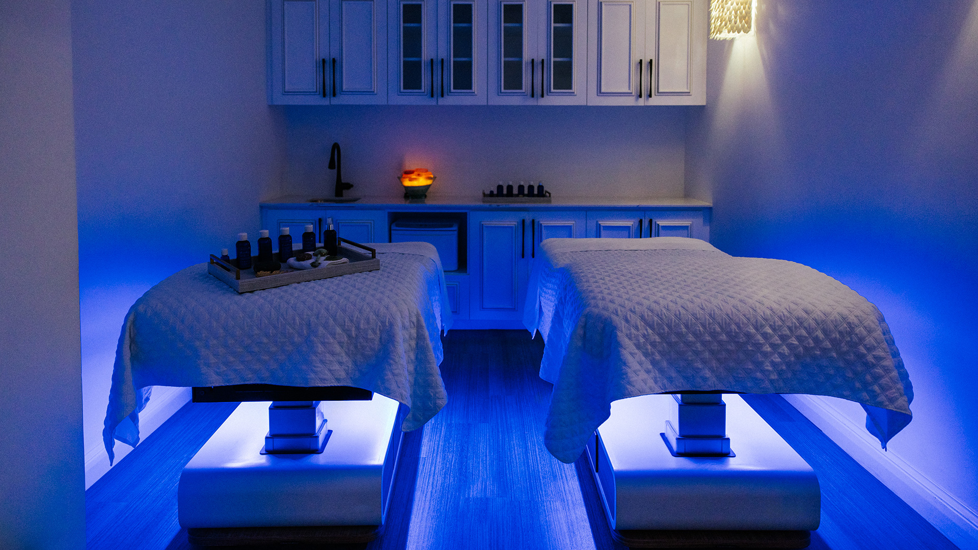 Two massage tables with white quilts are in a dimly lit spa room, glowing with blue accent lights. A tray with bottles sits on one table, and cabinets, a small sink, and candles are in the background.