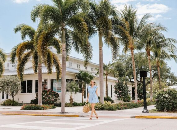 A woman in a light blue dress and sandals walks across a sunny street lined with palm trees, carrying a straw bag. Behind her is a white building surrounded by tropical landscaping. The sky is partly cloudy.