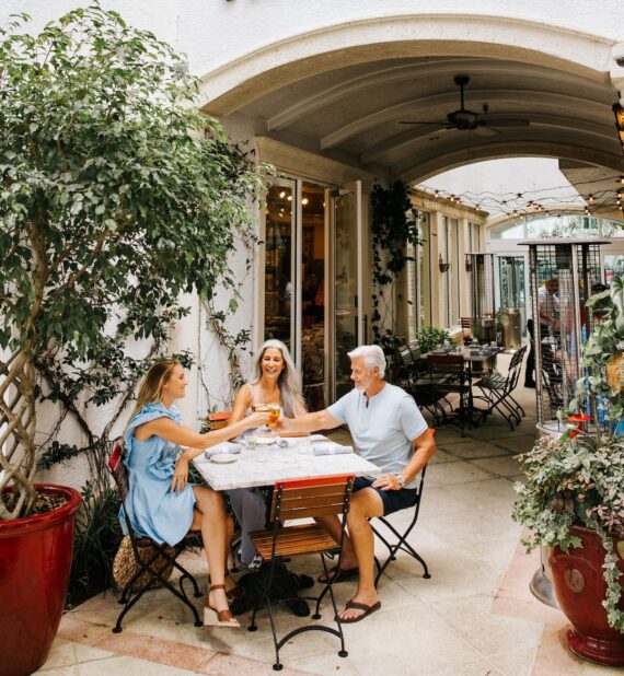 Three people sit at an outdoor cafe table under an archway, smiling and toasting drinks together. The setting is decorated with large potted plants and string lights, creating a cheerful, relaxed atmosphere.