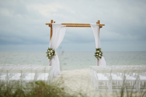 A wedding arch with white drapes and floral arrangements stands on a sandy beach, facing the ocean. Rows of white chairs are arranged for guests under a cloudy sky.