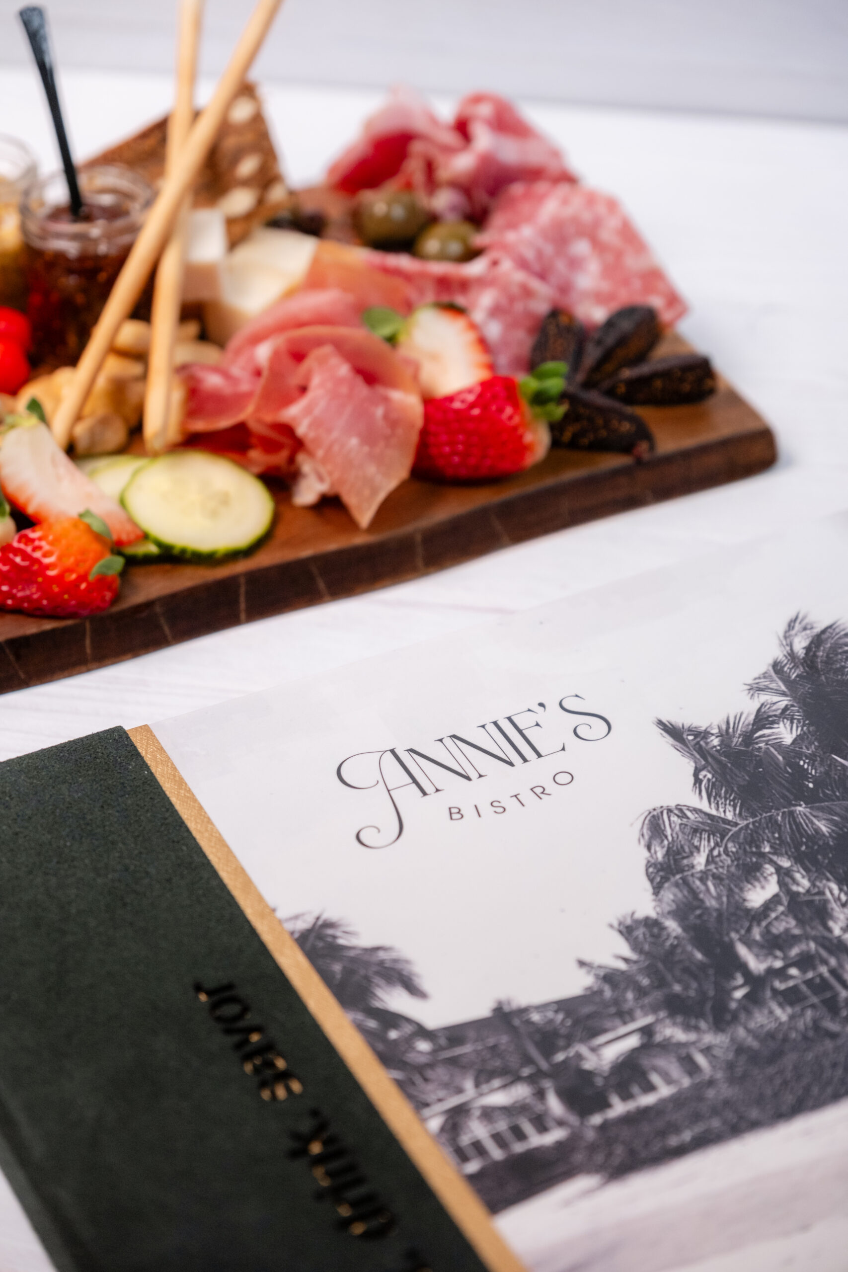 A charcuterie board with meats, cheeses, strawberries, and snacks sits behind menus from Annie’s Bistro, one of which features a black-and-white image of palm trees and a building.