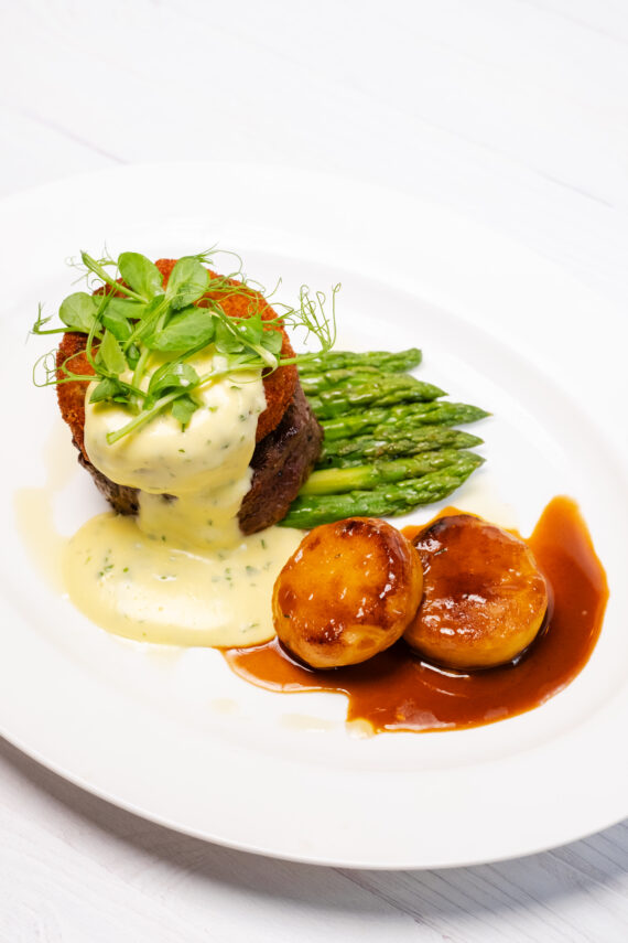 A plated gourmet meal featuring a beef filet topped with greens and hollandaise sauce, roasted potatoes with brown sauce, and a serving of asparagus spears, all arranged on a white plate.