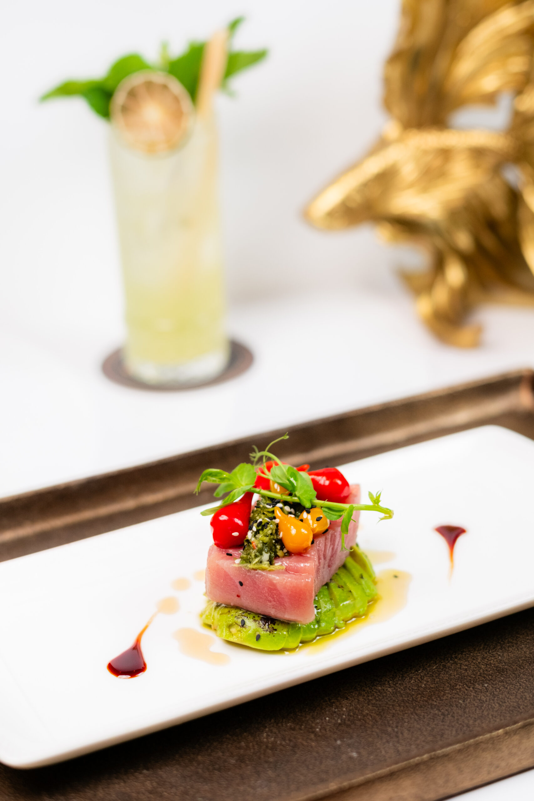 A gourmet dish of seared tuna on a bed of avocado, garnished with microgreens, cherry tomatoes, and sauce, is plated on a rectangular white dish. A cocktail with a garnish sits in the blurred background.