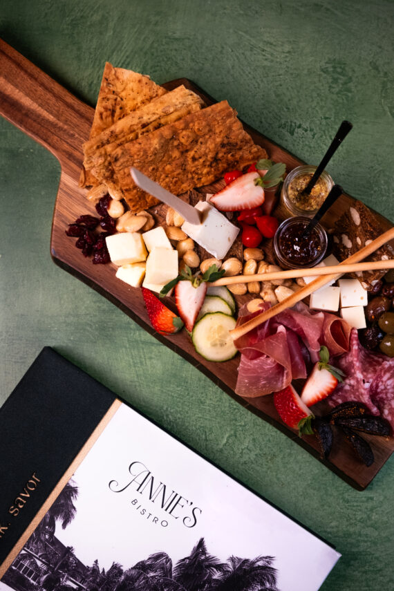 A wooden charcuterie board with assorted cheeses, crackers, nuts, strawberries, cured meats, olives, spreads, and fresh veggies, placed next to a bistro menu on a green surface.