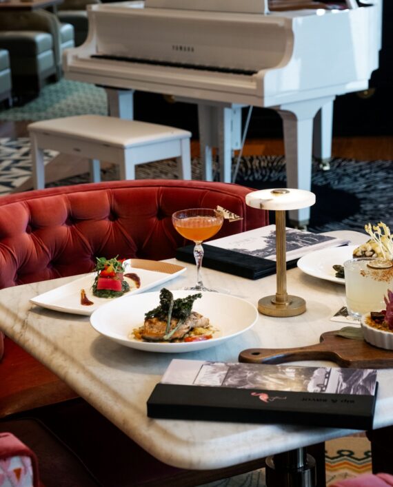 A marble table with elegant meals, cocktails, and menus sits beside a red velvet chair in a stylish restaurant; a white grand piano is in the background.