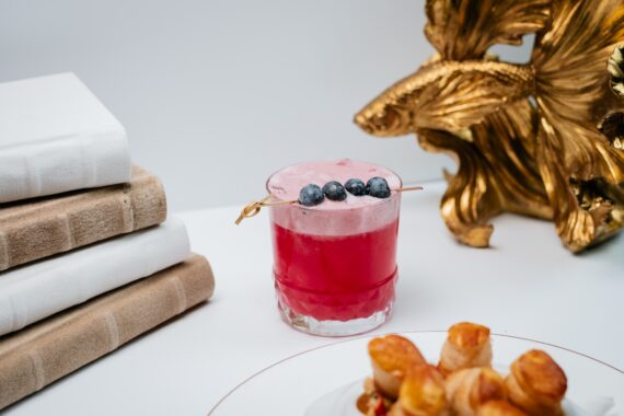 A pink cocktail garnished with three blueberries on a skewer sits on a white table next to stacked books and a decorative gold fish sculpture, with a plate of pastries partially visible in the foreground.