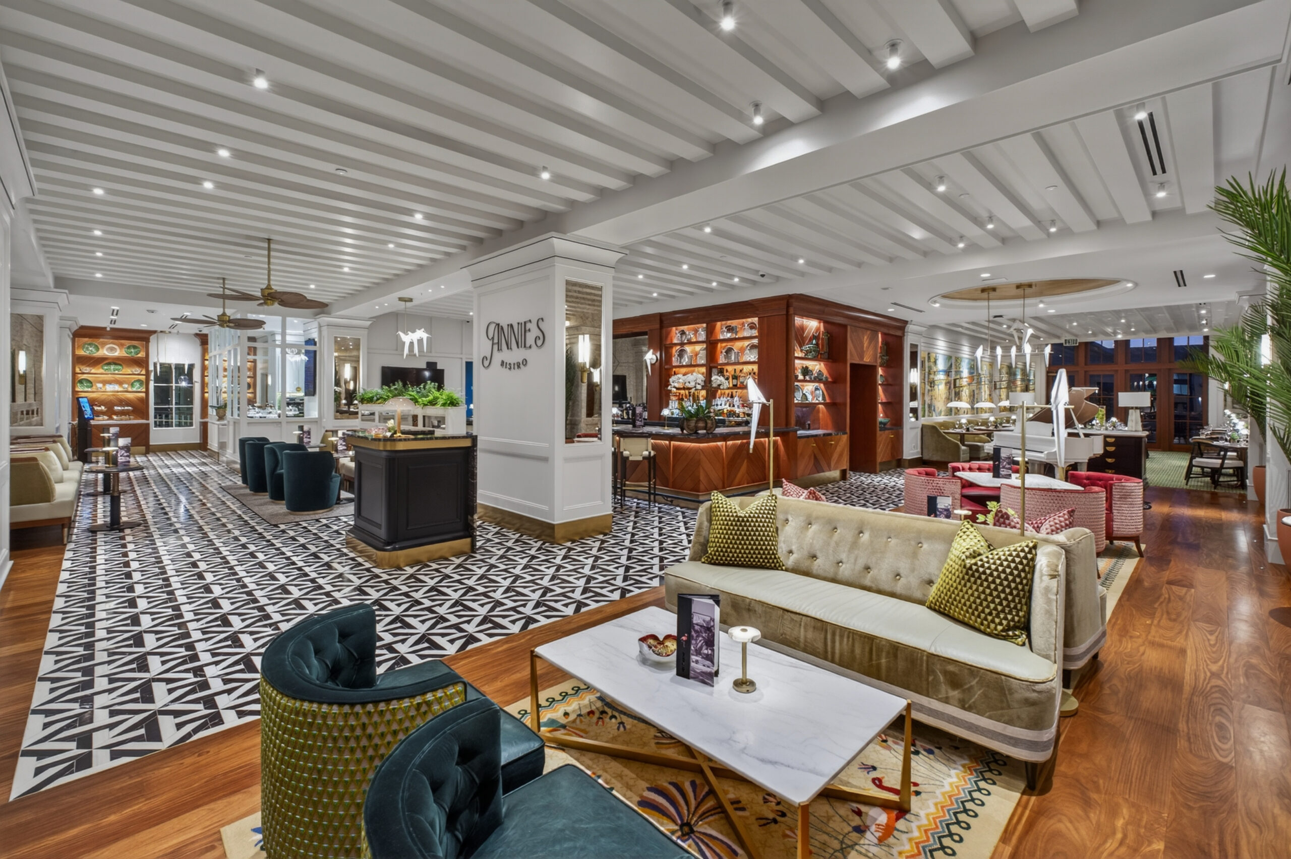 Spacious, modern hotel lobby with patterned tile and wood floors, stylish seating areas, bookshelves, and bright lighting. Cream sofas, green and gold chairs, and decorative pillows create a luxurious, inviting atmosphere.