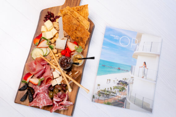 A charcuterie board with meats, cheeses, crackers, fruits, and dips beside a magazine showing a beach scene with a woman on a balcony and the text The OP, Issue #11, 2023 on a white surface.