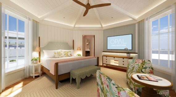 Bright, airy bedroom with large windows, a king-sized bed, floral-patterned chairs, a green bench, a ceiling fan, and a TV displaying Opal Collection on a dresser; coastal decor and soft natural light fill the space.