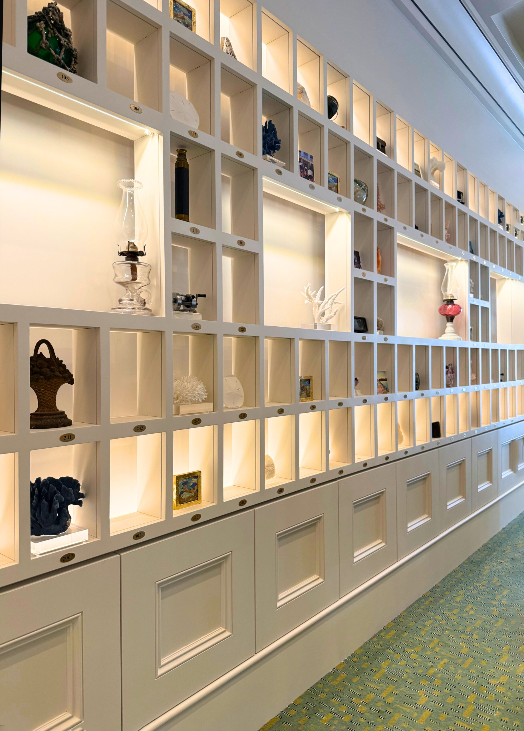 A wall of white, backlit display shelves filled with various decorative objects and collectibles, including glassware, sculptures, vases, and small art pieces, against a patterned green carpeted floor.