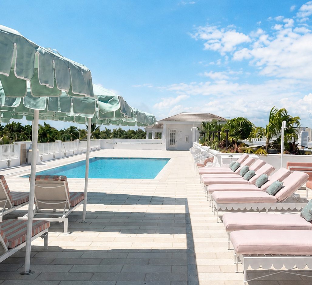 A rooftop swimming pool with light pink lounge chairs and mint green umbrellas on a sunny day, surrounded by white railings and palm trees, under a blue sky with scattered clouds.