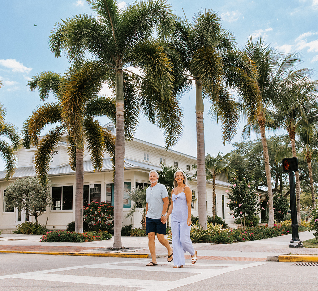 A smiling couple walks hand-in-hand across a palm tree-lined street on a sunny day, with a white building and lush landscaping in the background.