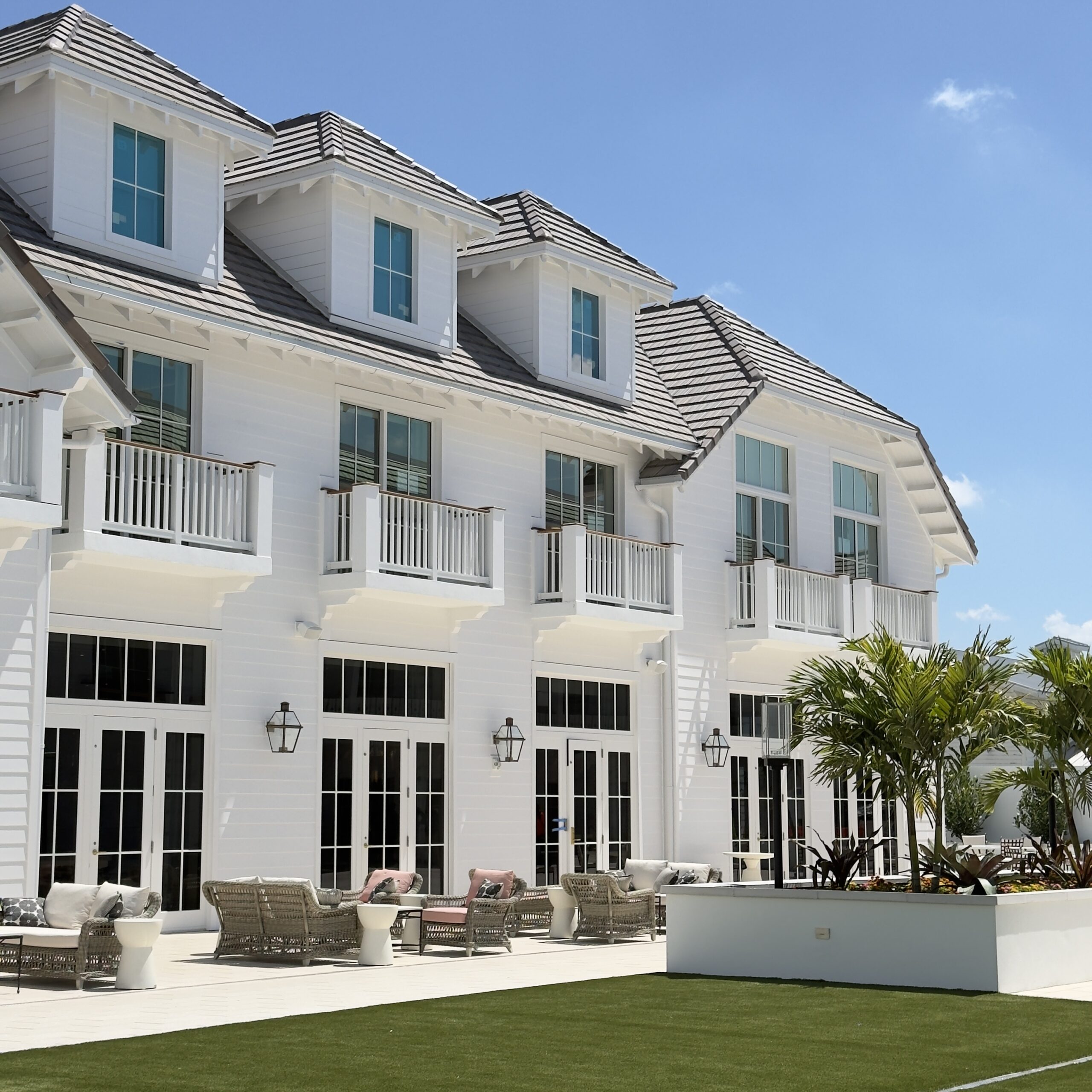 A large modern white house with multiple balconies, large windows, outdoor seating areas, and a manicured lawn, under a clear blue sky. Palm trees and patio furniture decorate the outdoor space.