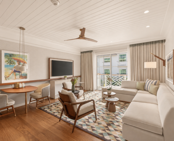 A bright, modern living room with a cream sectional sofa, geometric rug, round dining table with chairs, wall-mounted TV, ceiling fan, and large window with curtains overlooking a balcony and other buildings.