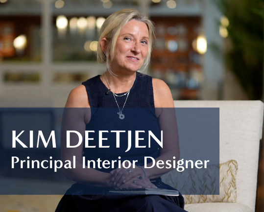A woman with blonde hair, wearing a sleeveless dark top, sits on a light-colored couch and smiles. Text overlay reads: Kim Deetjen, Principal Interior Designer. A blurred interior background is visible.