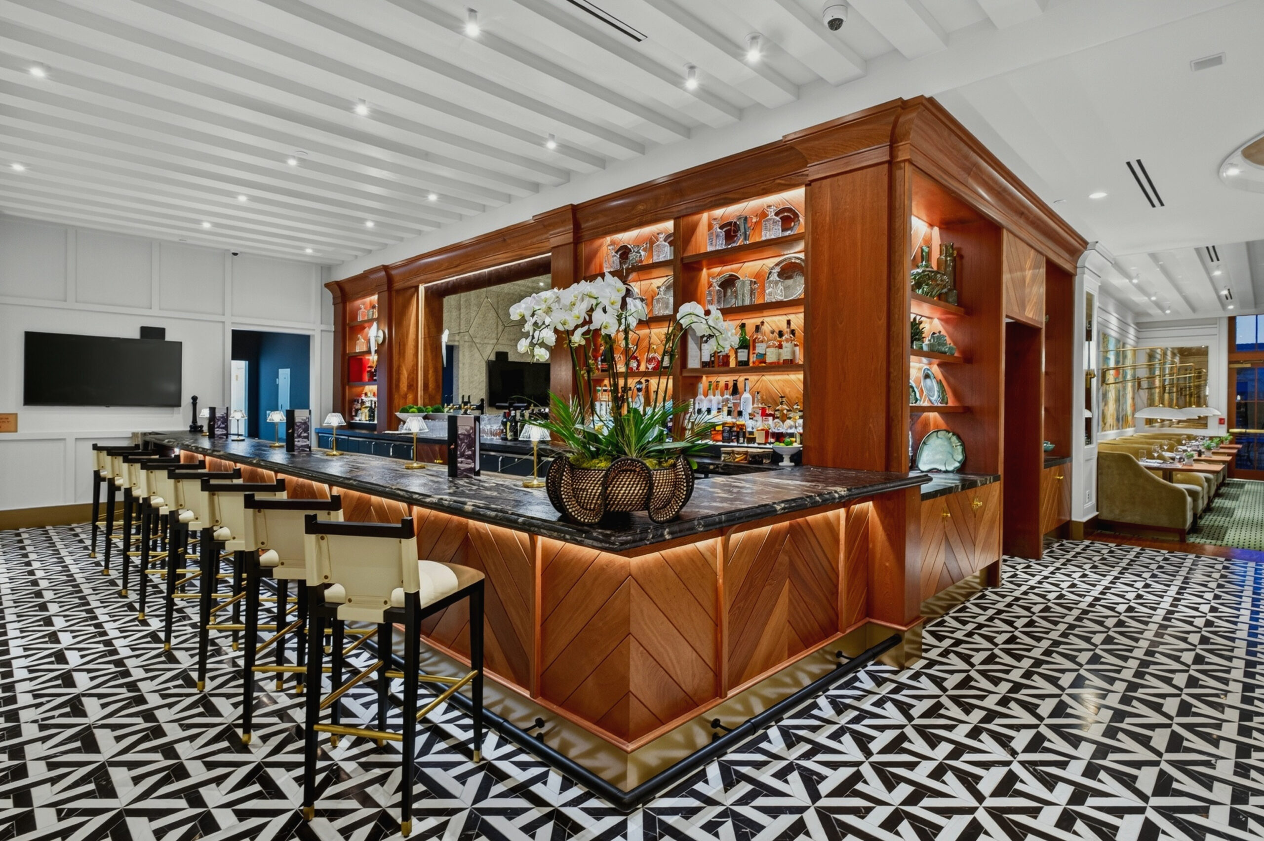 Elegant hotel bar with black-and-white patterned floor, marble countertop, cream barstools, wood paneling, backlit shelves with bottles and decor, and a central floral arrangement. Tables and chairs are visible in the background.