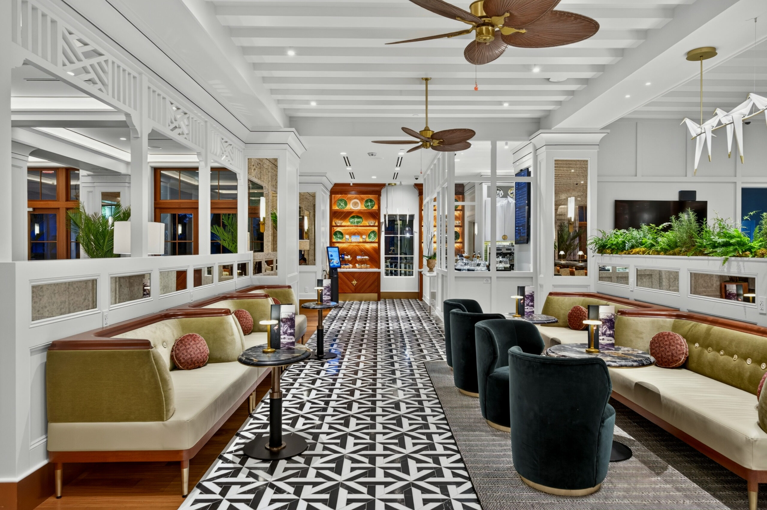 A stylish, modern lounge with patterned floors, green and beige sofas, black velvet chairs, ceiling fans, plants, and decorative shelves. The space is well-lit with large windows and white architectural accents.